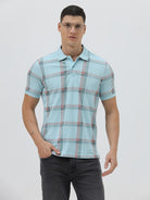 A man wearing a light blue short-sleeve polo shirt with a red and gray plaid pattern, paired with dark gray jeans, standing against a light gray studio background.