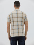 A man wearing a beige short-sleeve polo shirt with a brown and light blue plaid pattern, paired with dark blue jeans, standing against a light gray studio background.