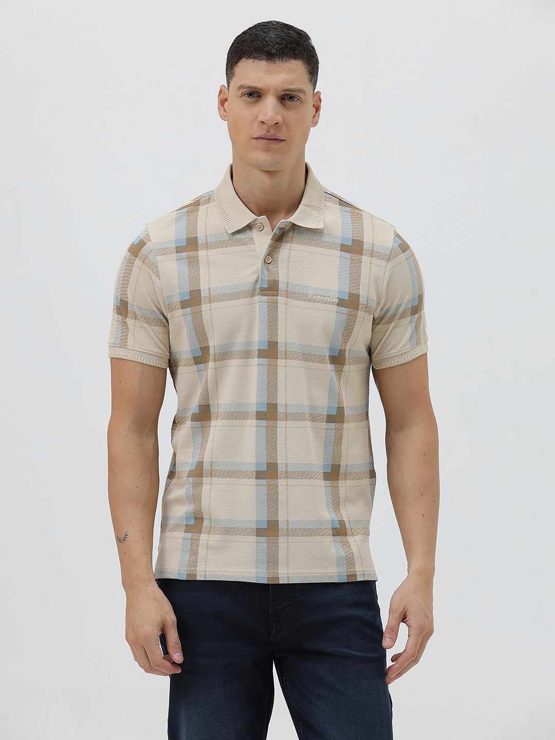 A man wearing a beige short-sleeve polo shirt with a brown and light blue plaid pattern, paired with dark blue jeans, standing against a light gray studio background.