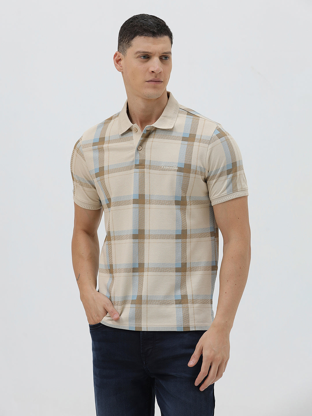A man wearing a beige short-sleeve polo shirt with a brown and light blue plaid pattern, paired with dark blue jeans, standing against a light gray studio background.