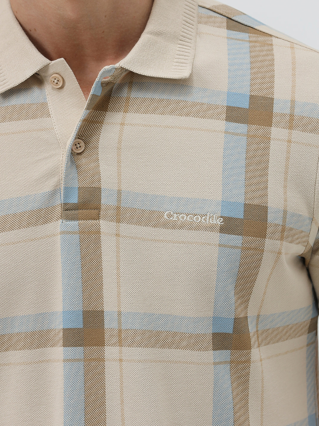A man wearing a beige short-sleeve polo shirt with a brown and light blue plaid pattern, paired with dark blue jeans, standing against a light gray studio background.