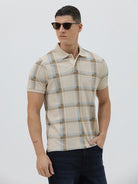 A man wearing a beige short-sleeve polo shirt with a brown and light blue plaid pattern, paired with dark blue jeans, standing against a light gray studio background.
