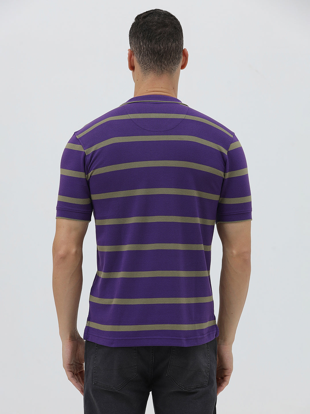 A man wearing a purple short-sleeve polo shirt with horizontal olive green stripes, paired with dark gray pants, standing against a light gray studio background.