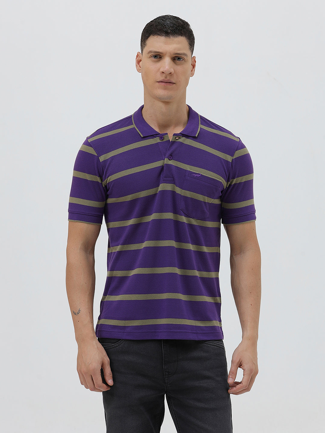 A man wearing a purple short-sleeve polo shirt with horizontal olive green stripes, paired with dark gray pants, standing against a light gray studio background.
