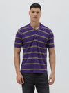 A man wearing a purple short-sleeve polo shirt with horizontal olive green stripes, paired with dark gray pants, standing against a light gray studio background.