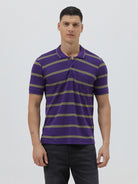 A man wearing a purple short-sleeve polo shirt with horizontal olive green stripes, paired with dark gray pants, standing against a light gray studio background.