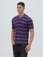 A man wearing a purple short-sleeve polo shirt with horizontal olive green stripes, paired with dark gray pants, standing against a light gray studio background.