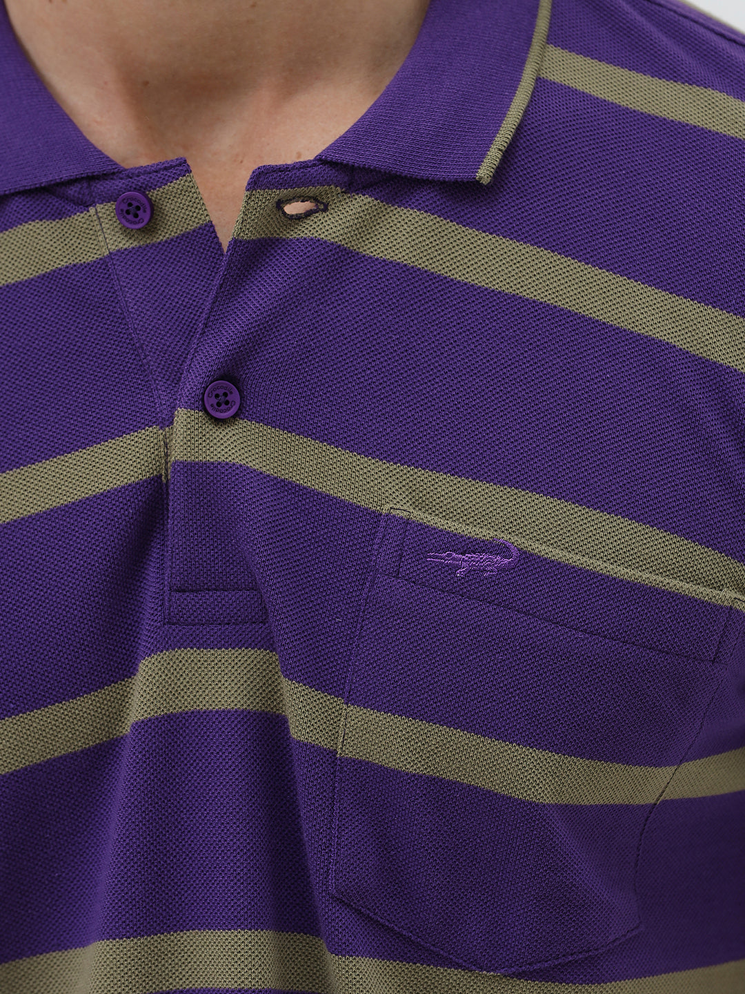 A man wearing a purple short-sleeve polo shirt with horizontal olive green stripes, paired with dark gray pants, standing against a light gray studio background.