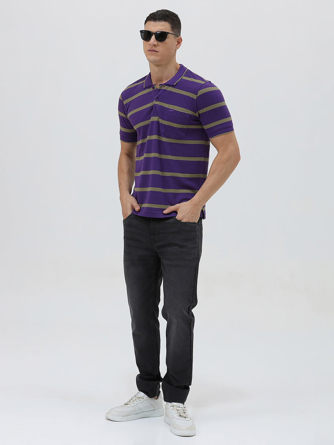 A man wearing a purple short-sleeve polo shirt with horizontal olive green stripes, paired with dark gray pants, standing against a light gray studio background.