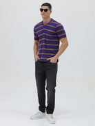 A man wearing a purple short-sleeve polo shirt with horizontal olive green stripes, paired with dark gray pants, standing against a light gray studio background.