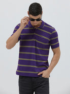 A man wearing a purple short-sleeve polo shirt with horizontal olive green stripes, paired with dark gray pants, standing against a light gray studio background.