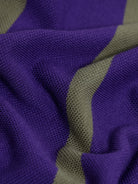A man wearing a purple short-sleeve polo shirt with horizontal olive green stripes, paired with dark gray pants, standing against a light gray studio background.