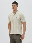 A man wearing a light beige short-sleeve polo shirt with subtle horizontal stripe details and a small crocodile logo on the left chest, paired with olive green trousers, against a plain light gray background.