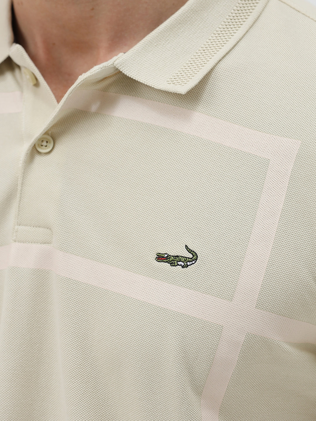 A man wearing a light beige short-sleeve polo shirt with subtle horizontal stripe details and a small crocodile logo on the left chest, paired with olive green trousers, against a plain light gray background.