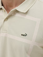 A man wearing a light beige short-sleeve polo shirt with subtle horizontal stripe details and a small crocodile logo on the left chest, paired with olive green trousers, against a plain light gray background.