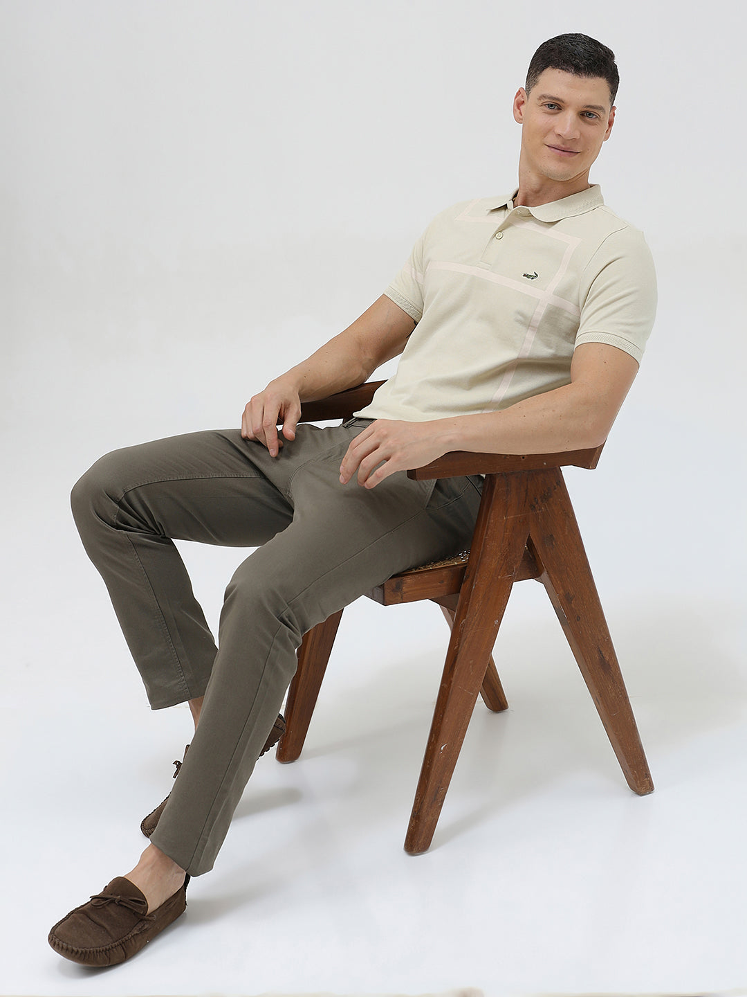 A man wearing a light beige short-sleeve polo shirt with subtle horizontal stripe details and a small crocodile logo on the left chest, paired with olive green trousers, against a plain light gray background.