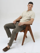 A man wearing a light beige short-sleeve polo shirt with subtle horizontal stripe details and a small crocodile logo on the left chest, paired with olive green trousers, against a plain light gray background.