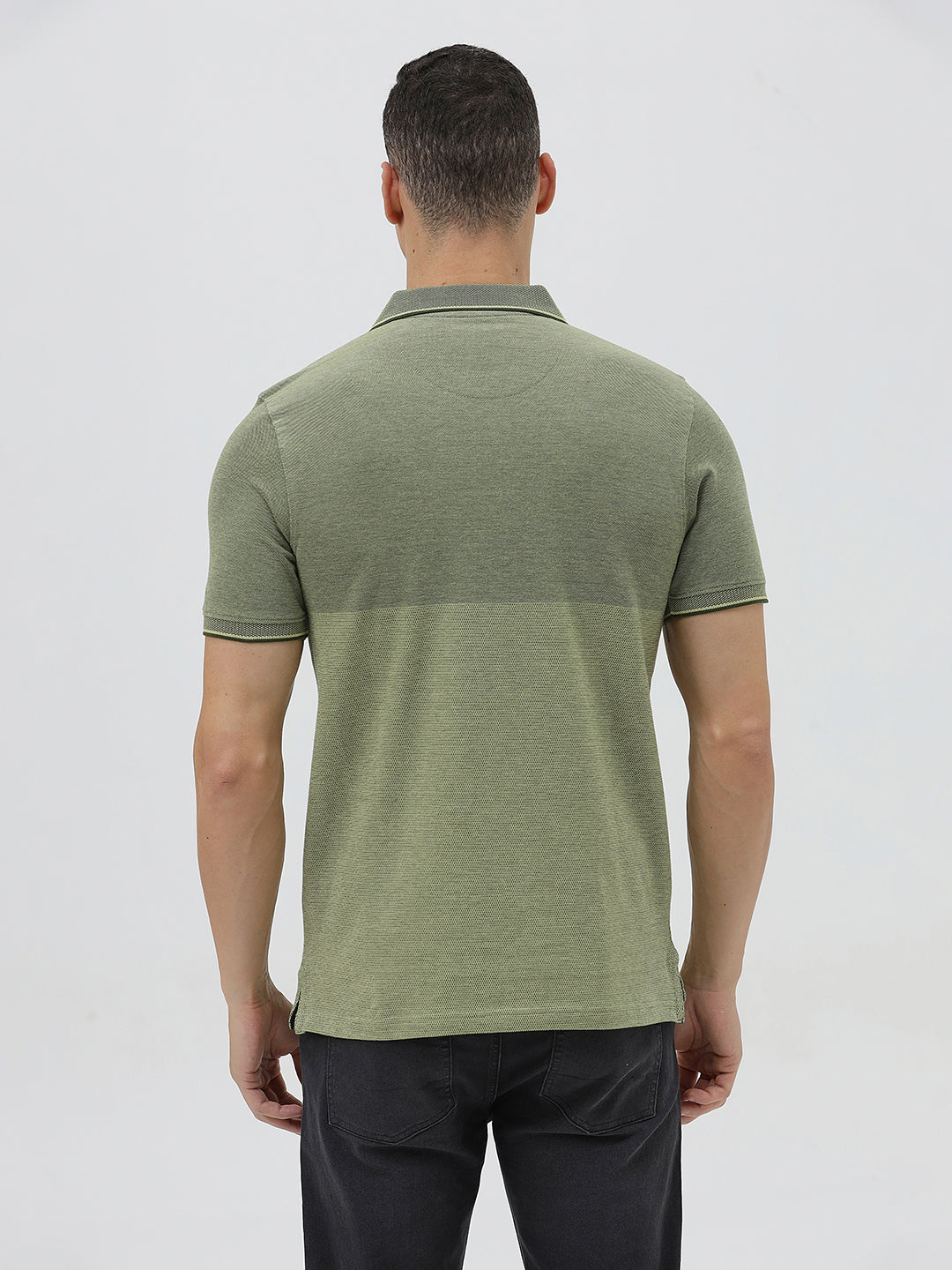 A man wearing a short-sleeve polo shirt in olive green with a subtle horizontal color-blocked stripe and a small gold crest logo on the left chest, paired with dark pants, against a light gray studio background.