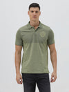 A man wearing a short-sleeve polo shirt in olive green with a subtle horizontal color-blocked stripe and a small gold crest logo on the left chest, paired with dark pants, against a light gray studio background.