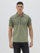A man wearing a short-sleeve polo shirt in olive green with a subtle horizontal color-blocked stripe and a small gold crest logo on the left chest, paired with dark pants, against a light gray studio background.
