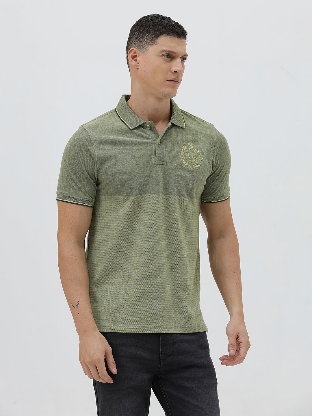 A man wearing a short-sleeve polo shirt in olive green with a subtle horizontal color-blocked stripe and a small gold crest logo on the left chest, paired with dark pants, against a light gray studio background.