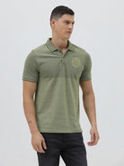 A man wearing a short-sleeve polo shirt in olive green with a subtle horizontal color-blocked stripe and a small gold crest logo on the left chest, paired with dark pants, against a light gray studio background.