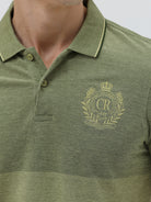 A man wearing a short-sleeve polo shirt in olive green with a subtle horizontal color-blocked stripe and a small gold crest logo on the left chest, paired with dark pants, against a light gray studio background.