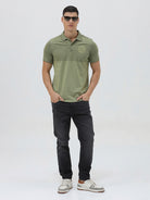 A man wearing a short-sleeve polo shirt in olive green with a subtle horizontal color-blocked stripe and a small gold crest logo on the left chest, paired with dark pants, against a light gray studio background.