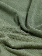 A man wearing a short-sleeve polo shirt in olive green with a subtle horizontal color-blocked stripe and a small gold crest logo on the left chest, paired with dark pants, against a light gray studio background.
