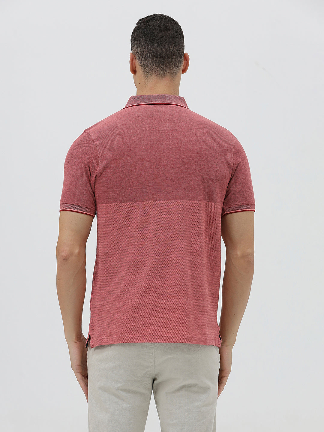 A man wearing a heathered dusty rose pink polo shirt with a tonal embroidered crest on the left chest, paired with light beige trousers, against a plain light gray background.