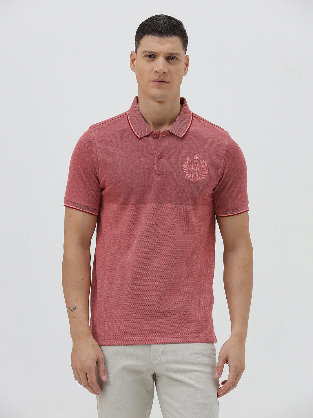 A man wearing a heathered dusty rose pink polo shirt with a tonal embroidered crest on the left chest, paired with light beige trousers, against a plain light gray background.