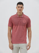 A man wearing a heathered dusty rose pink polo shirt with a tonal embroidered crest on the left chest, paired with light beige trousers, against a plain light gray background.