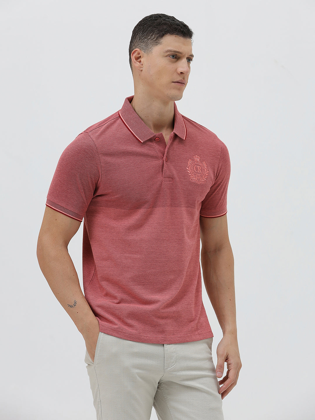 A man wearing a heathered dusty rose pink polo shirt with a tonal embroidered crest on the left chest, paired with light beige trousers, against a plain light gray background.