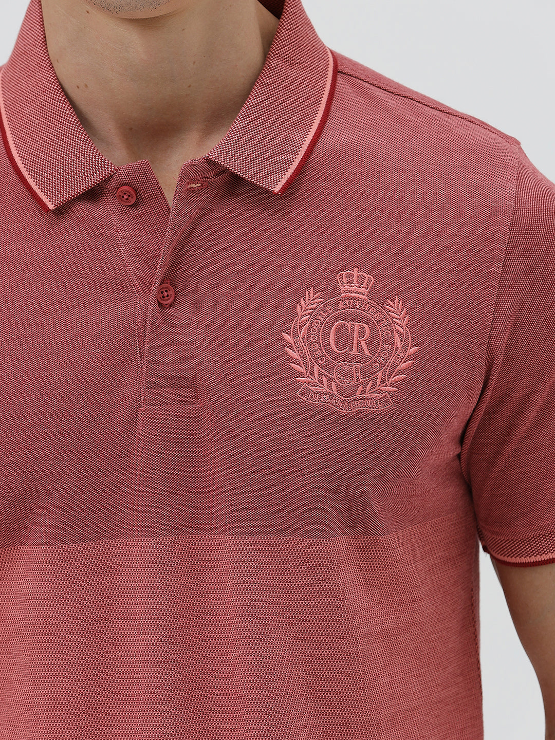 A man wearing a heathered dusty rose pink polo shirt with a tonal embroidered crest on the left chest, paired with light beige trousers, against a plain light gray background.