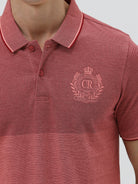 A man wearing a heathered dusty rose pink polo shirt with a tonal embroidered crest on the left chest, paired with light beige trousers, against a plain light gray background.