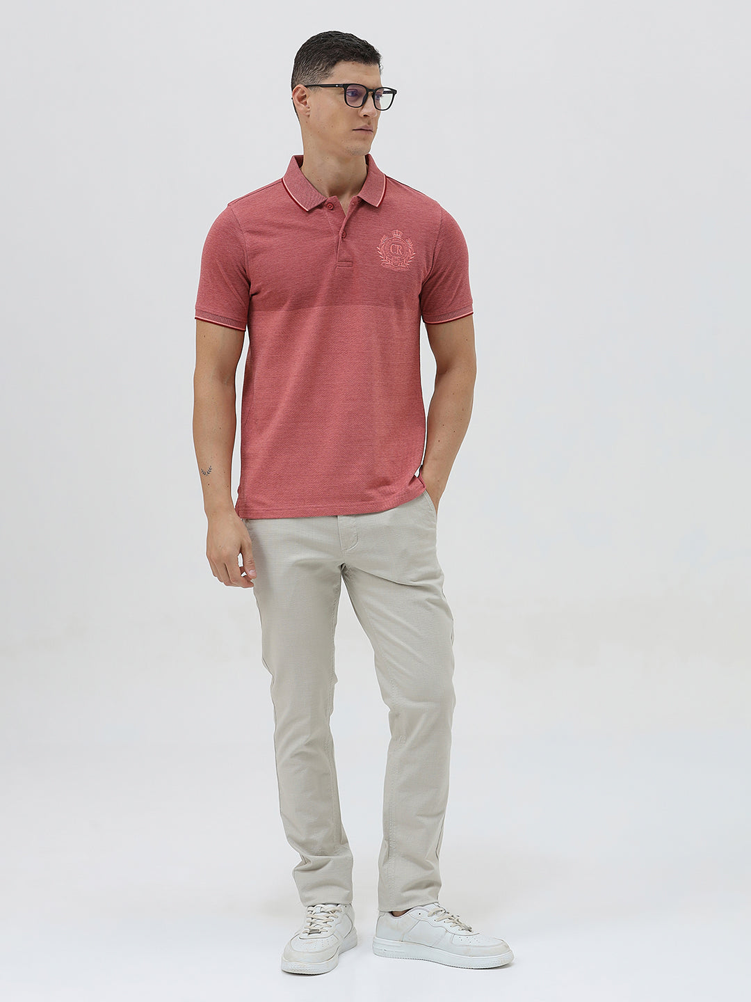 A man wearing a heathered dusty rose pink polo shirt with a tonal embroidered crest on the left chest, paired with light beige trousers, against a plain light gray background.
