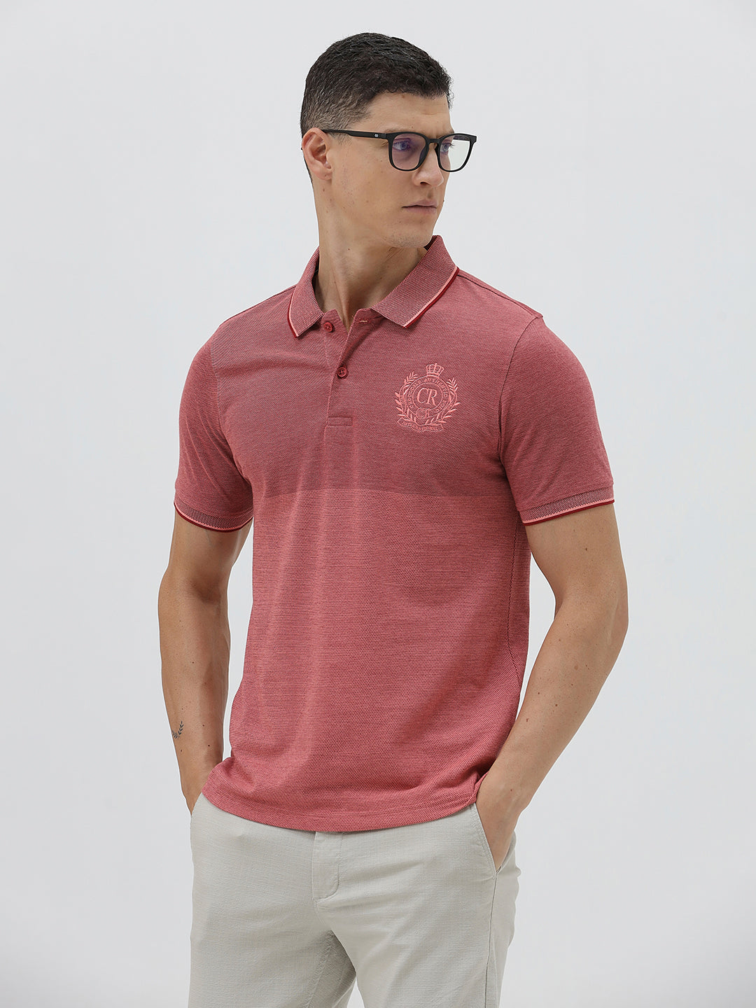 A man wearing a heathered dusty rose pink polo shirt with a tonal embroidered crest on the left chest, paired with light beige trousers, against a plain light gray background.