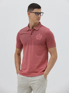 A man wearing a heathered dusty rose pink polo shirt with a tonal embroidered crest on the left chest, paired with light beige trousers, against a plain light gray background.