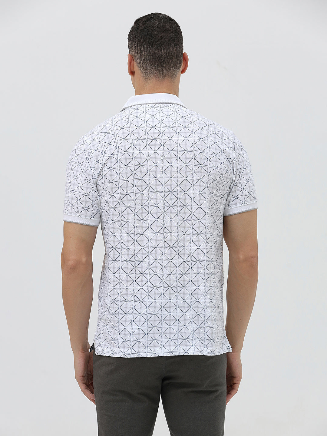 A man wearing a white short-sleeve polo shirt with a gray geometric pattern, paired with dark gray trousers, standing against a light gray studio background.