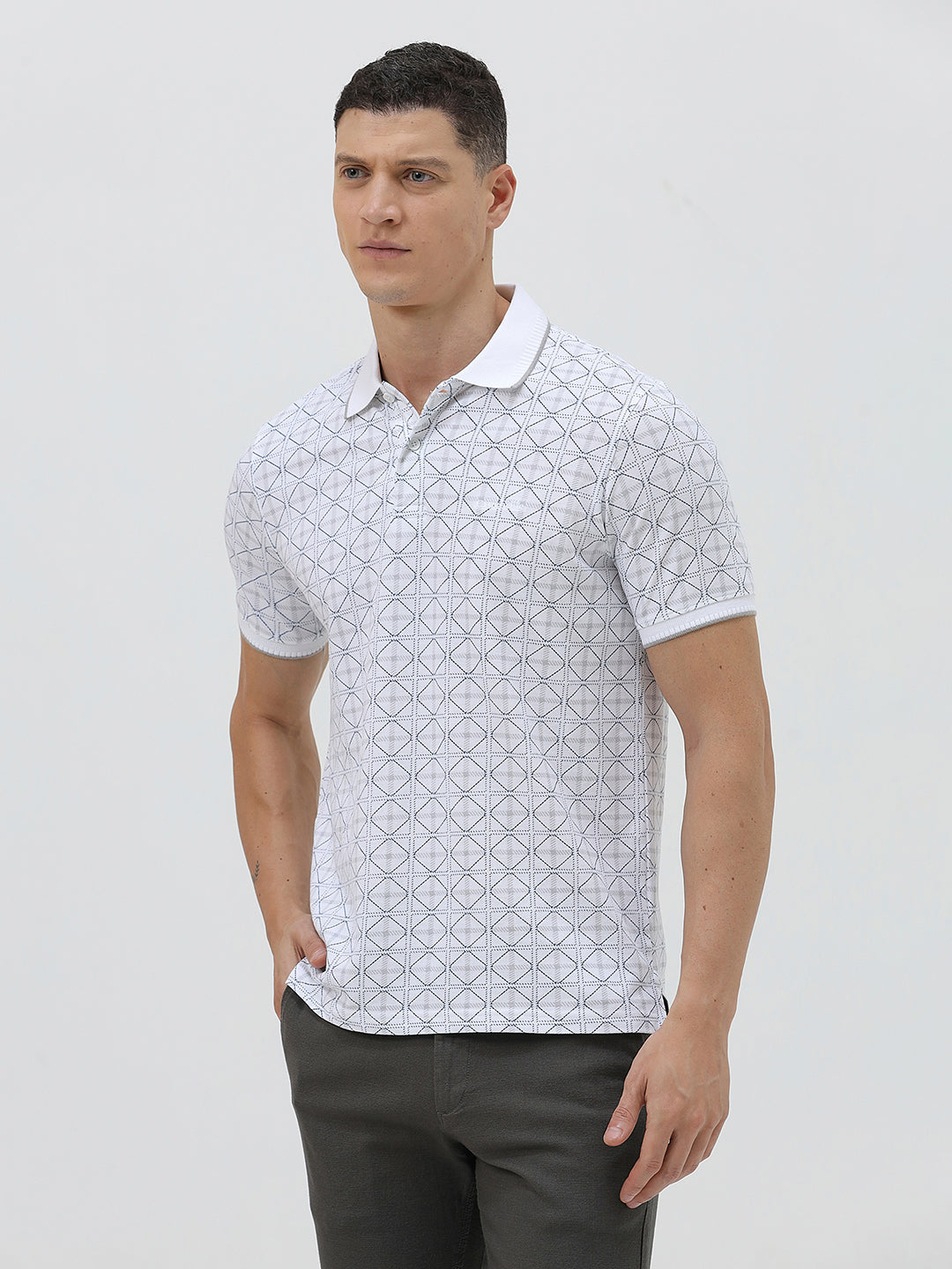 A man wearing a white short-sleeve polo shirt with a gray geometric pattern, paired with dark gray trousers, standing against a light gray studio background.