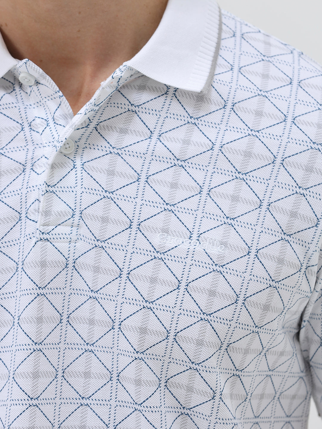 A man wearing a white short-sleeve polo shirt with a gray geometric pattern, paired with dark gray trousers, standing against a light gray studio background.