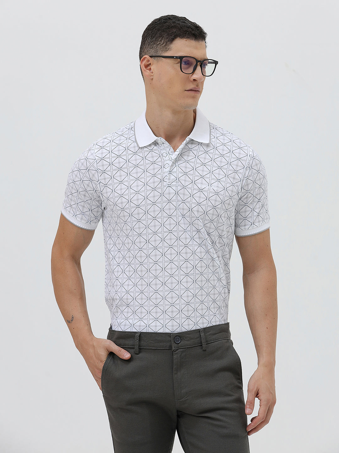 A man wearing a white short-sleeve polo shirt with a gray geometric pattern, paired with dark gray trousers, standing against a light gray studio background.