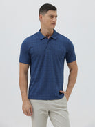 A man wearing a blue short-sleeve polo shirt with a subtle geometric pattern, paired with beige trousers, standing against a light gray studio background.
