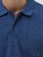 A man wearing a blue short-sleeve polo shirt with a subtle geometric pattern, paired with beige trousers, standing against a light gray studio background.