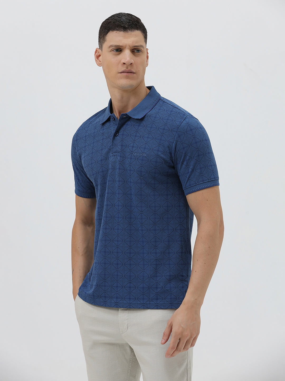A man wearing a blue short-sleeve polo shirt with a subtle geometric pattern, paired with beige trousers, standing against a light gray studio background.