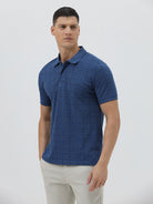 A man wearing a blue short-sleeve polo shirt with a subtle geometric pattern, paired with beige trousers, standing against a light gray studio background.