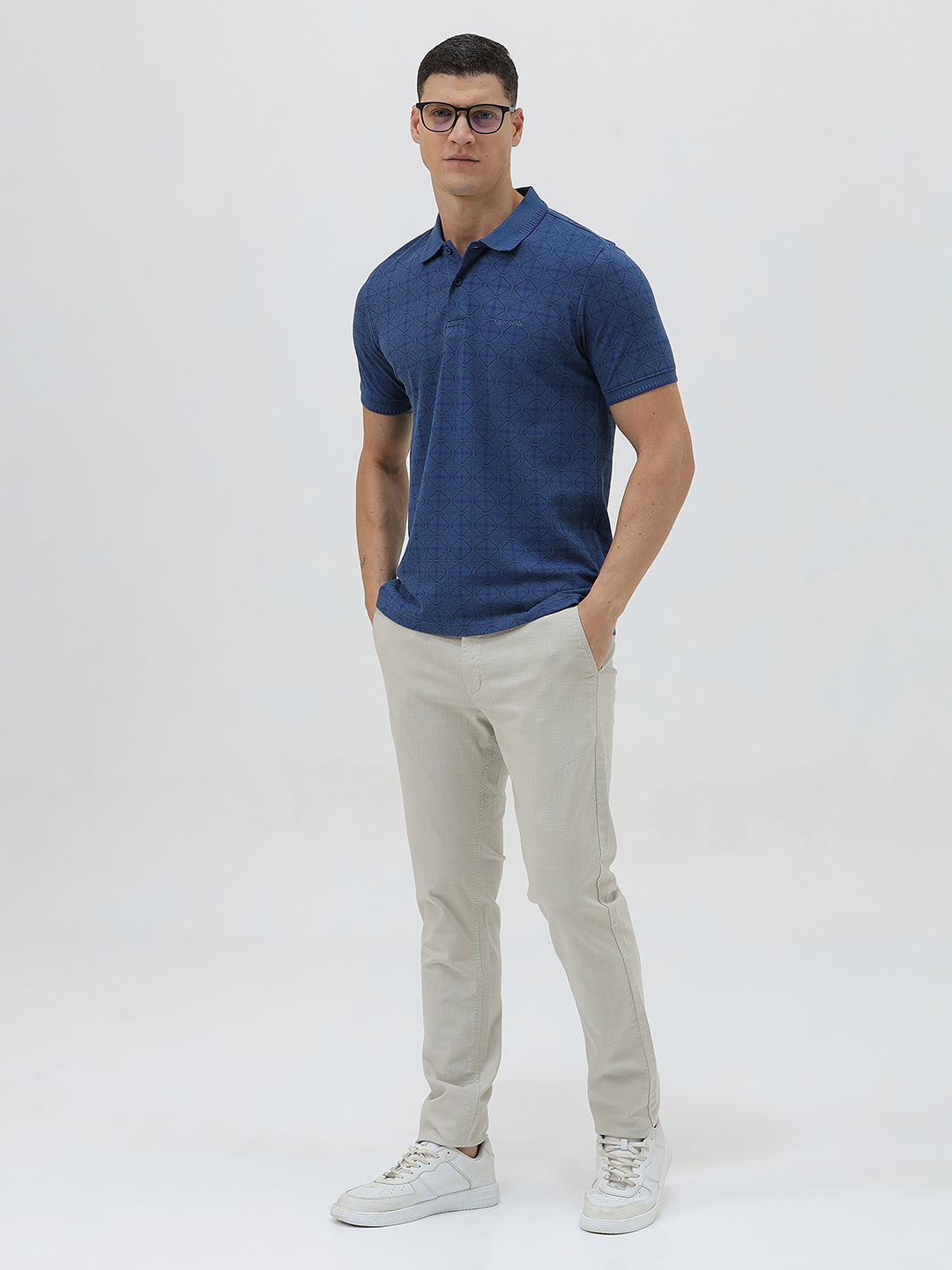 A man wearing a blue short-sleeve polo shirt with a subtle geometric pattern, paired with beige trousers, standing against a light gray studio background.