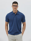 A man wearing a blue short-sleeve polo shirt with a subtle geometric pattern, paired with beige trousers, standing against a light gray studio background.