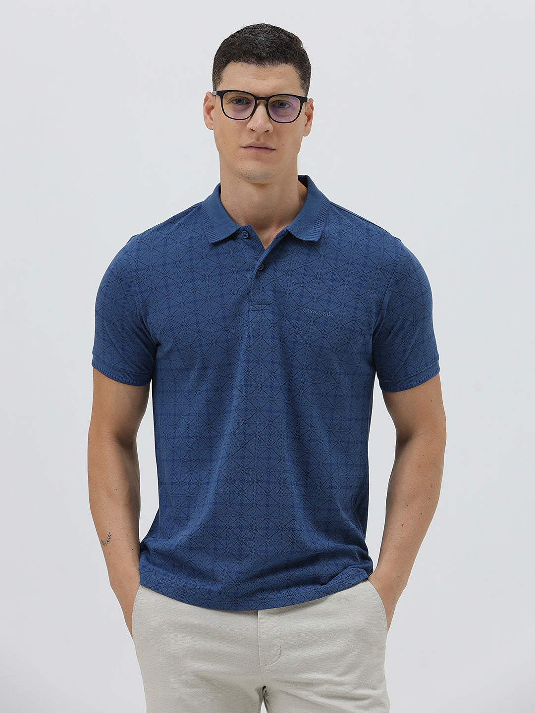 A man wearing a blue short-sleeve polo shirt with a subtle geometric pattern, paired with beige trousers, standing against a light gray studio background.