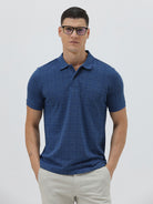 A man wearing a blue short-sleeve polo shirt with a subtle geometric pattern, paired with beige trousers, standing against a light gray studio background.